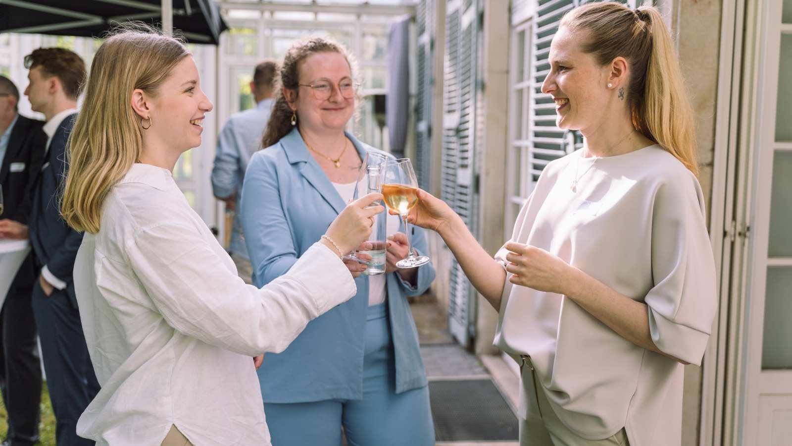 Drei Frauen stehen zusammen in einer sommerlichen Atmosphäre und stoßen miteinander an.